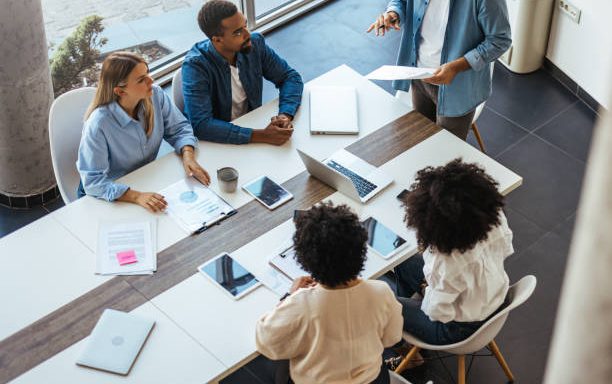 featured-image-1783051220 A diverse group of professionals in a modern office setting engaged in a collaborative business meeting. Team members are actively discussing strategies and projects with the aid of technology and paperwork.